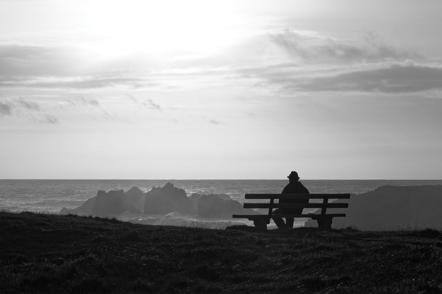 Silhouette of person sitting alone on bench overlooking mountainous horizon at sunset.