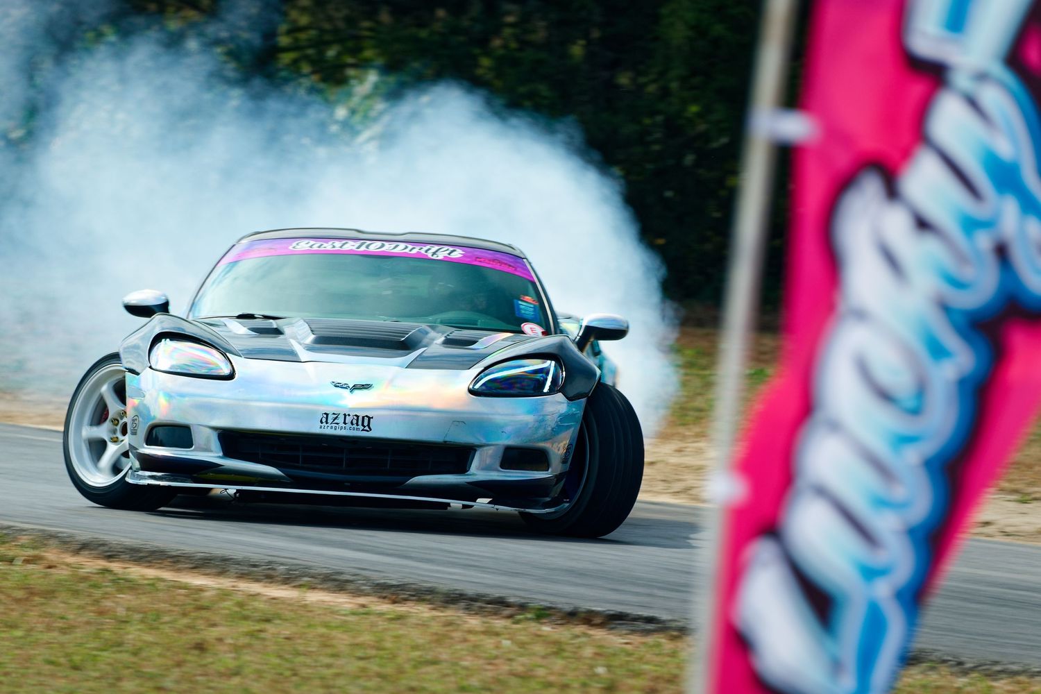 A Corvette drifting on racetrack with smoke trailing behind and pink banner in foreground.