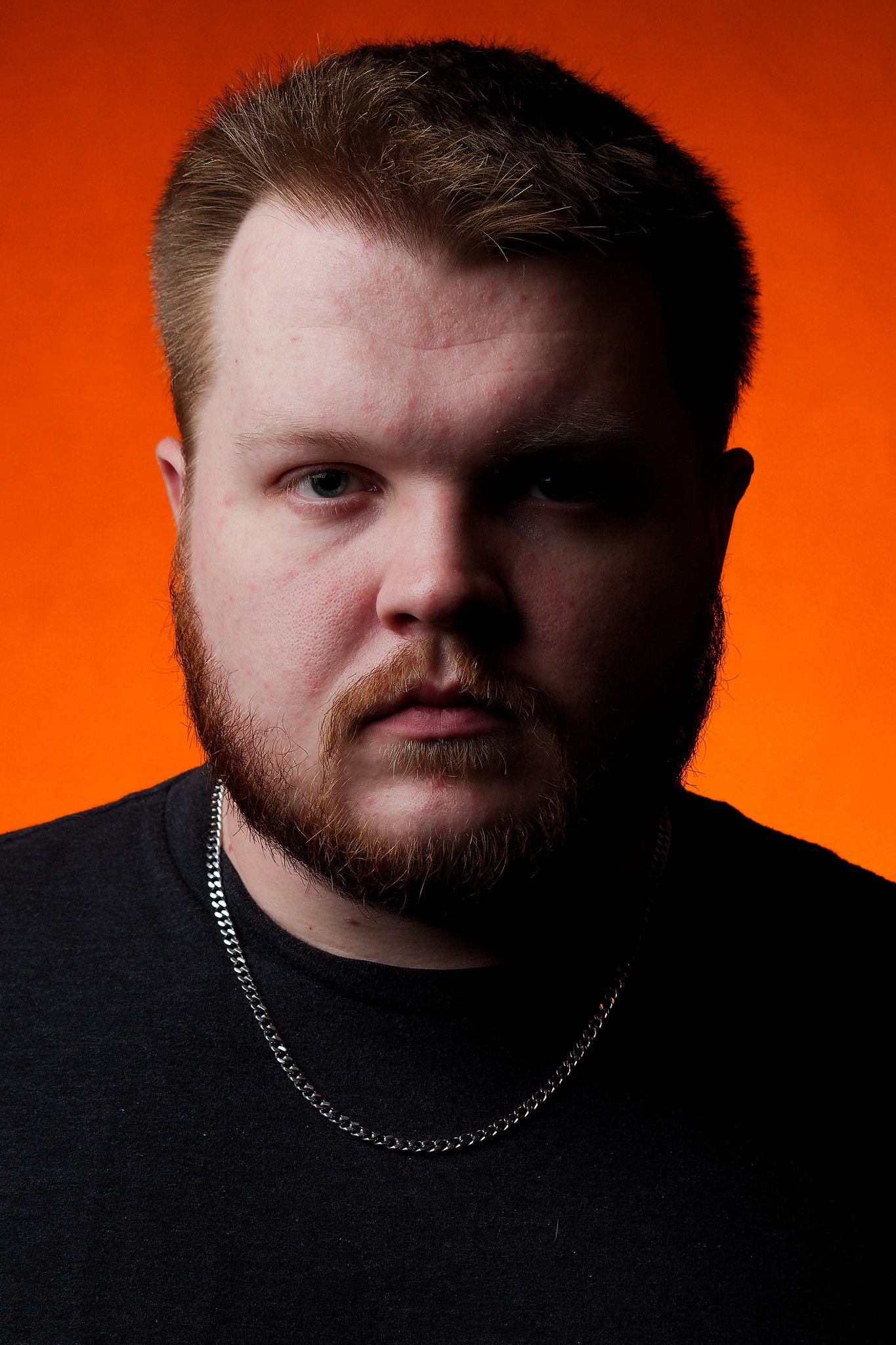 A portrait against an orange backdrop featuring a man in black clothing with a silver chain necklace.