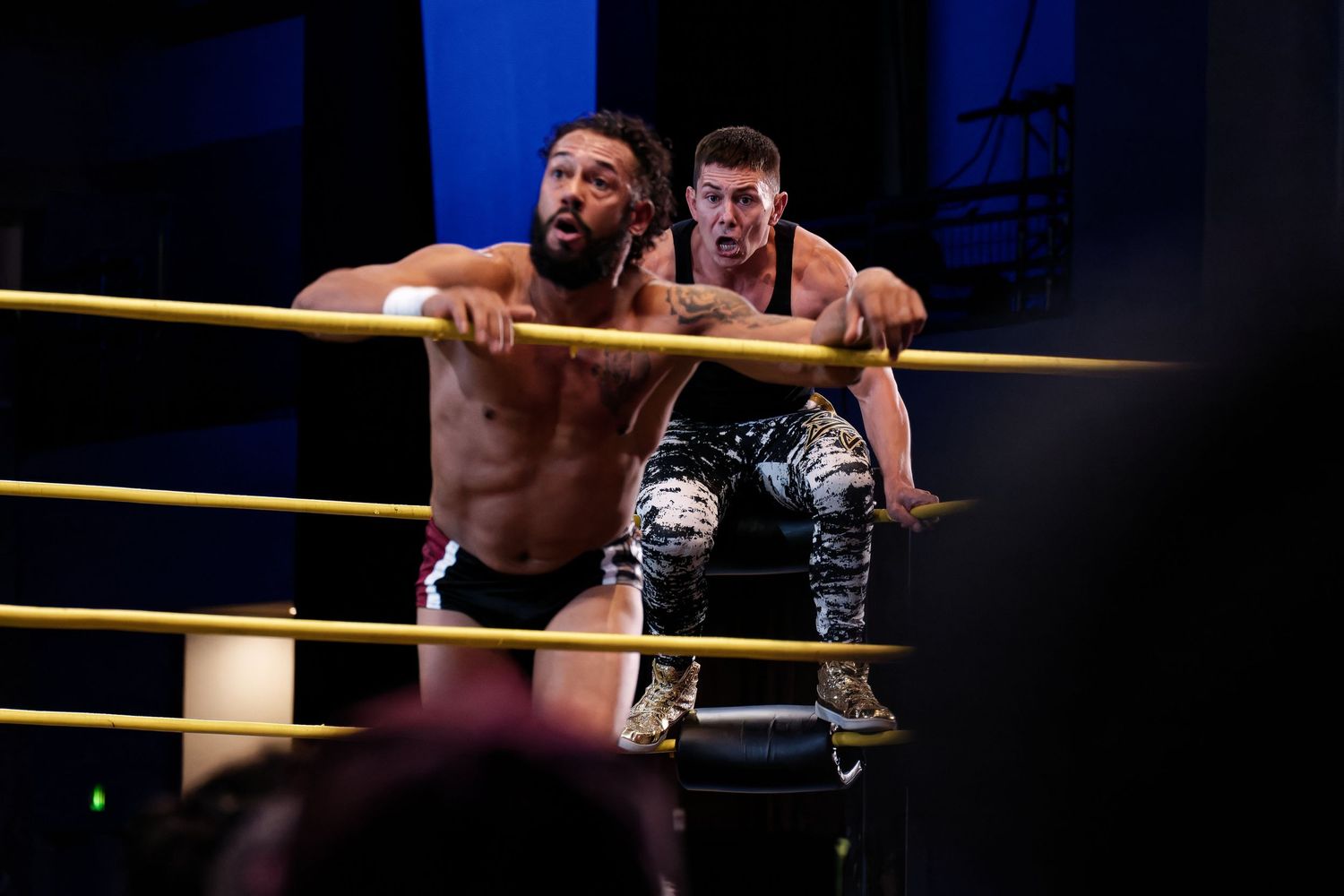 Two wrestlers competing in dramatic match on wrestling ring ropes.