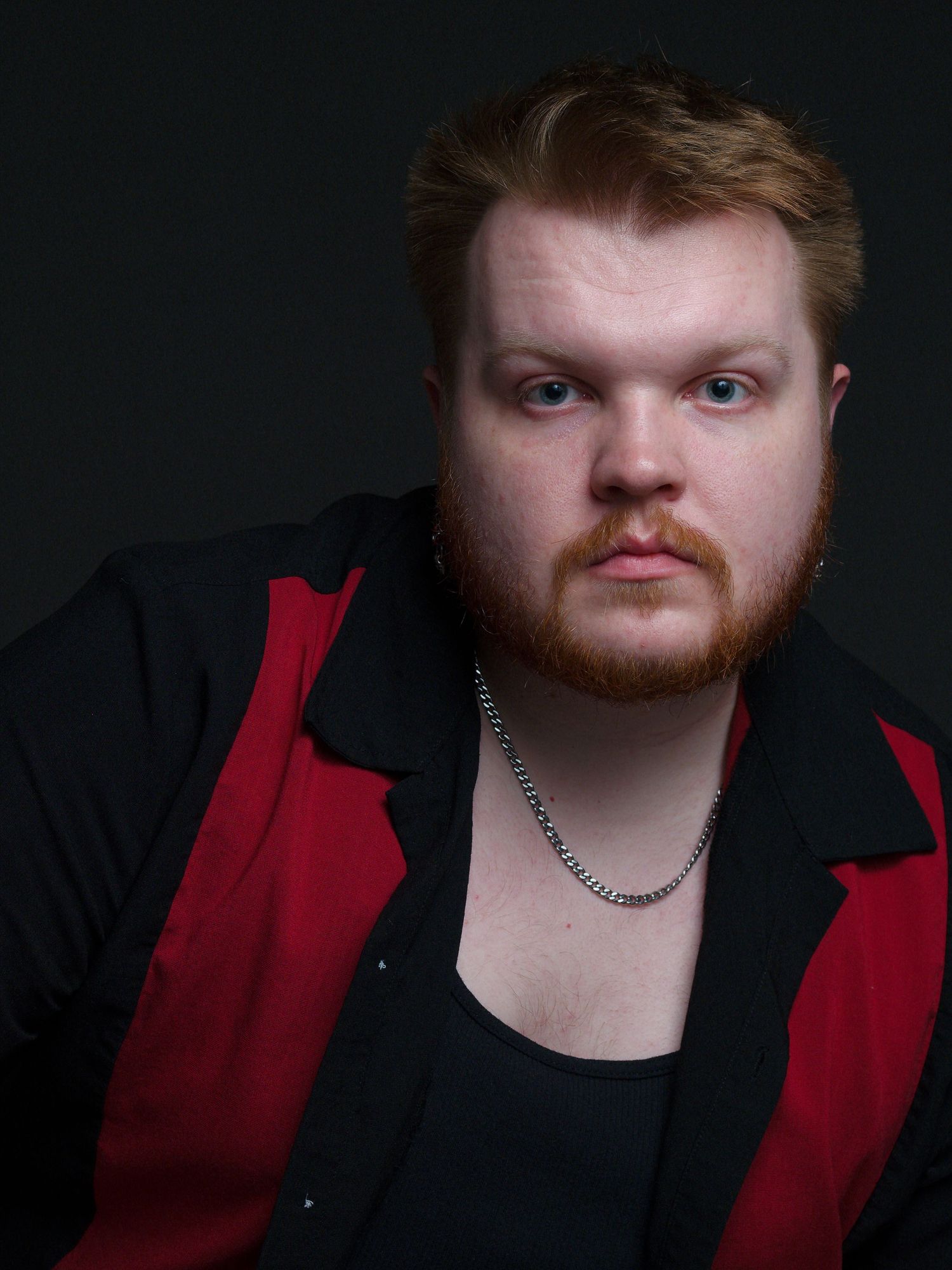 A portrait of a man wearing a red and black shirt and a silver chain. The background is black
