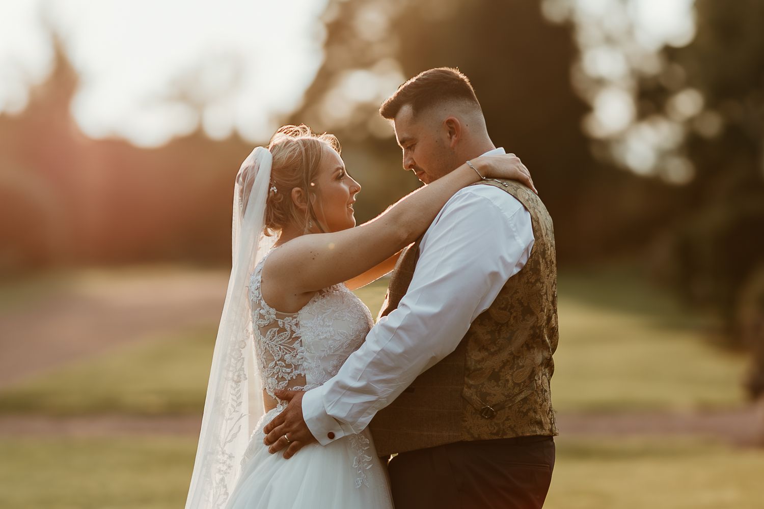 A couple in formal wedding attire share an intimate embrace during golden hour in a beautiful outdoor setting.