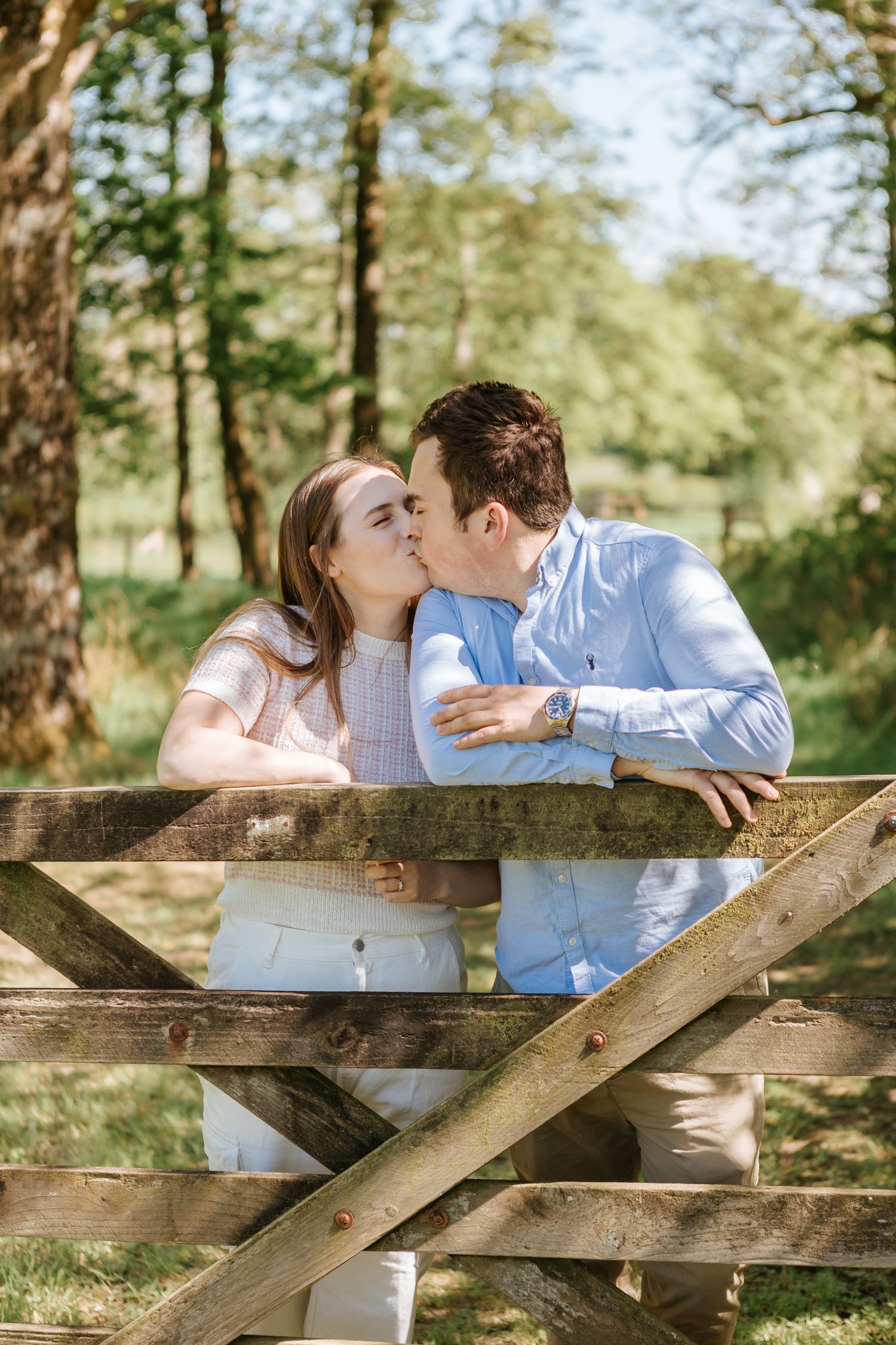 A romantic couple shares a sweet kiss while leaning on a rustic wooden gate in a sunlit forest setting. Alexandra Maxwell Photography