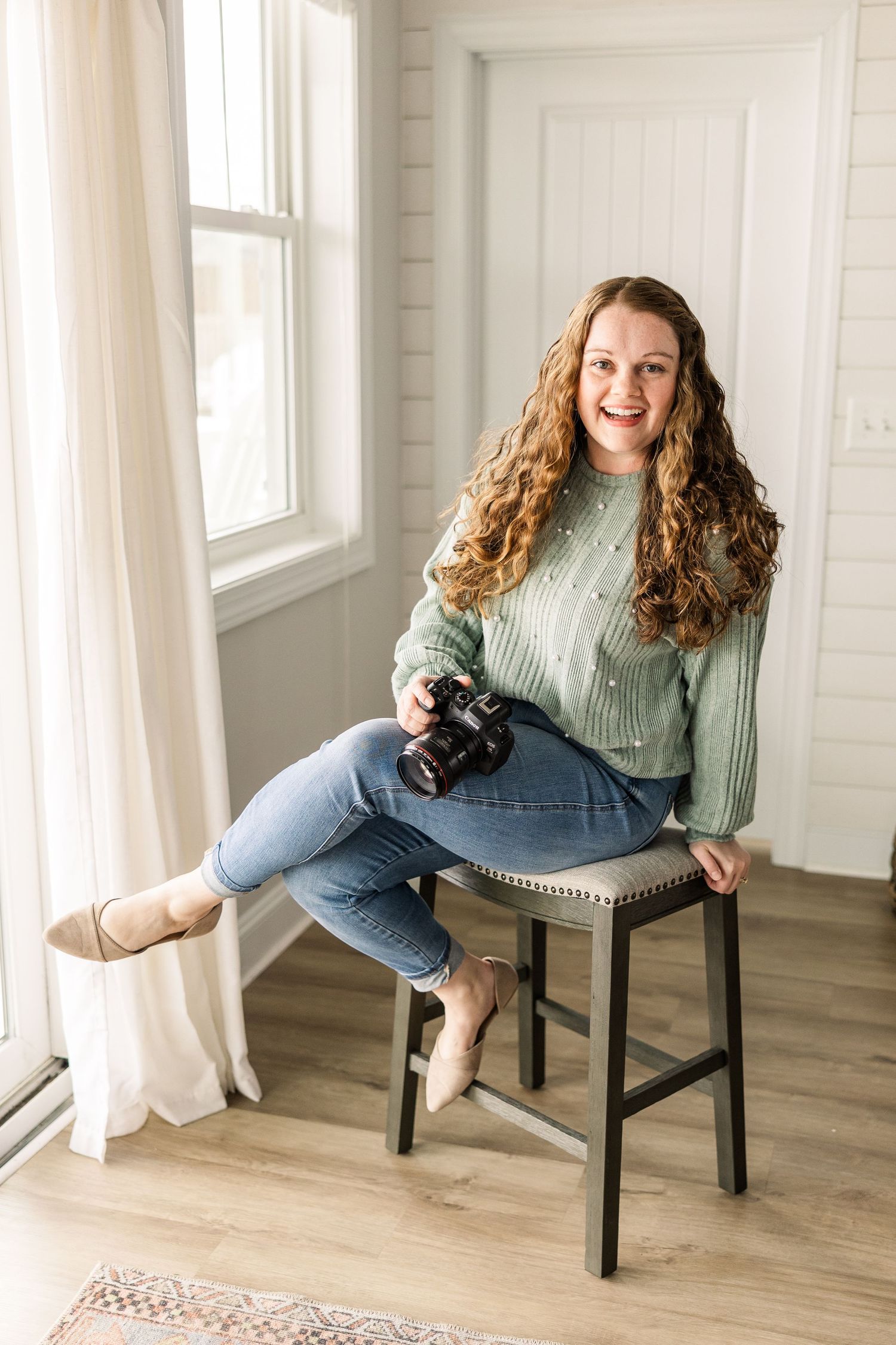 Photographer sits on wooden stool near window in bright white room holding camera while wearing mint sweater and jeans.