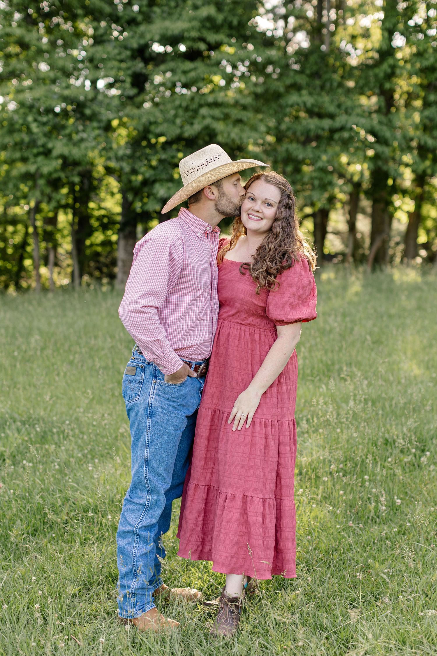 A couple in coordinating pink and red outfits poses together in a sunny green field with trees in the background.