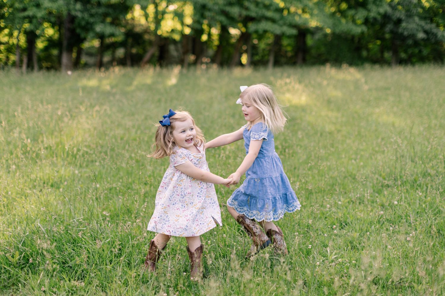 Two young children in dresses dancing and holding hands together in a sunny grassy field.