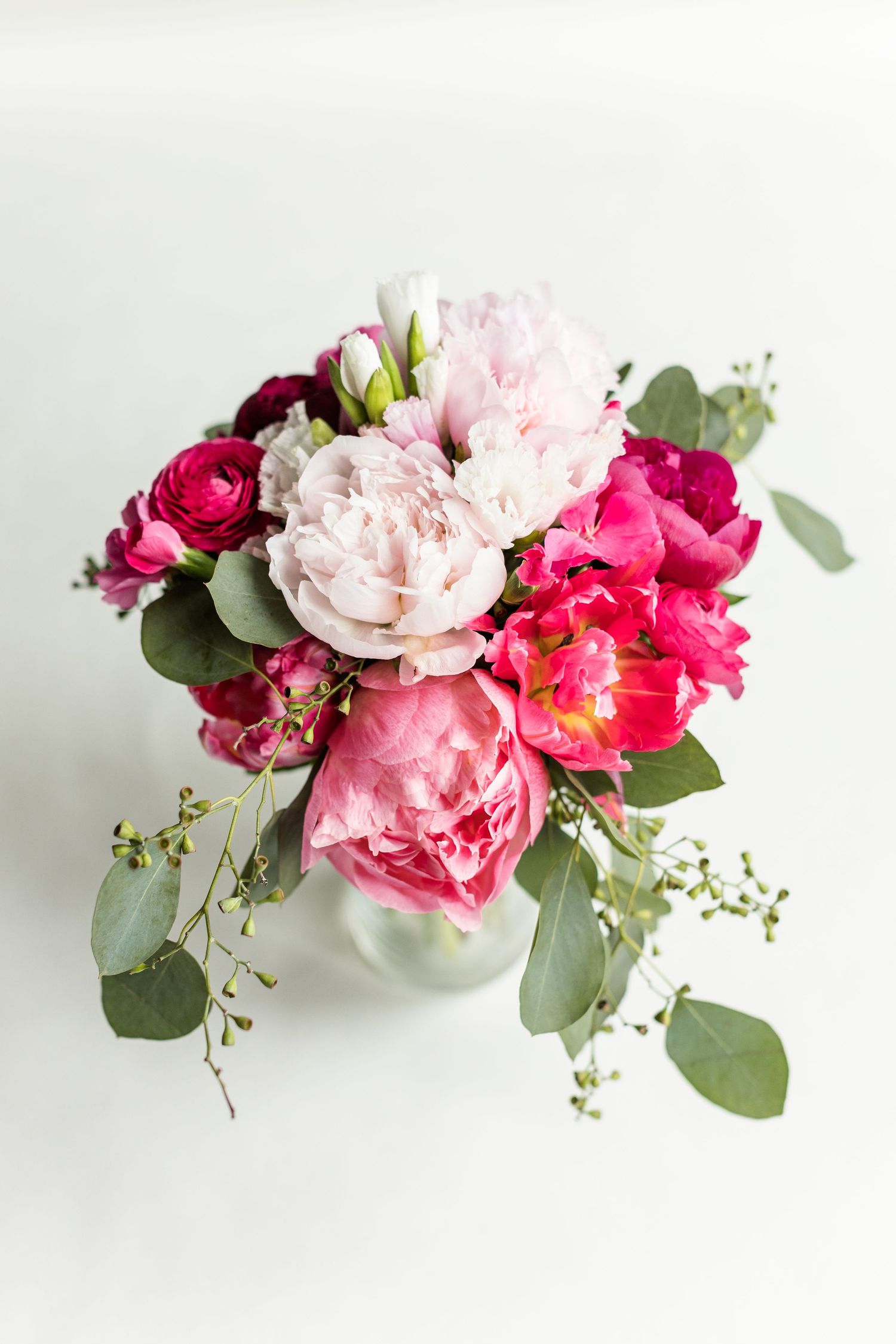 Beautiful floral arrangement with pink peonies, red ranunculus, and eucalyptus leaves in a white vase against light background.