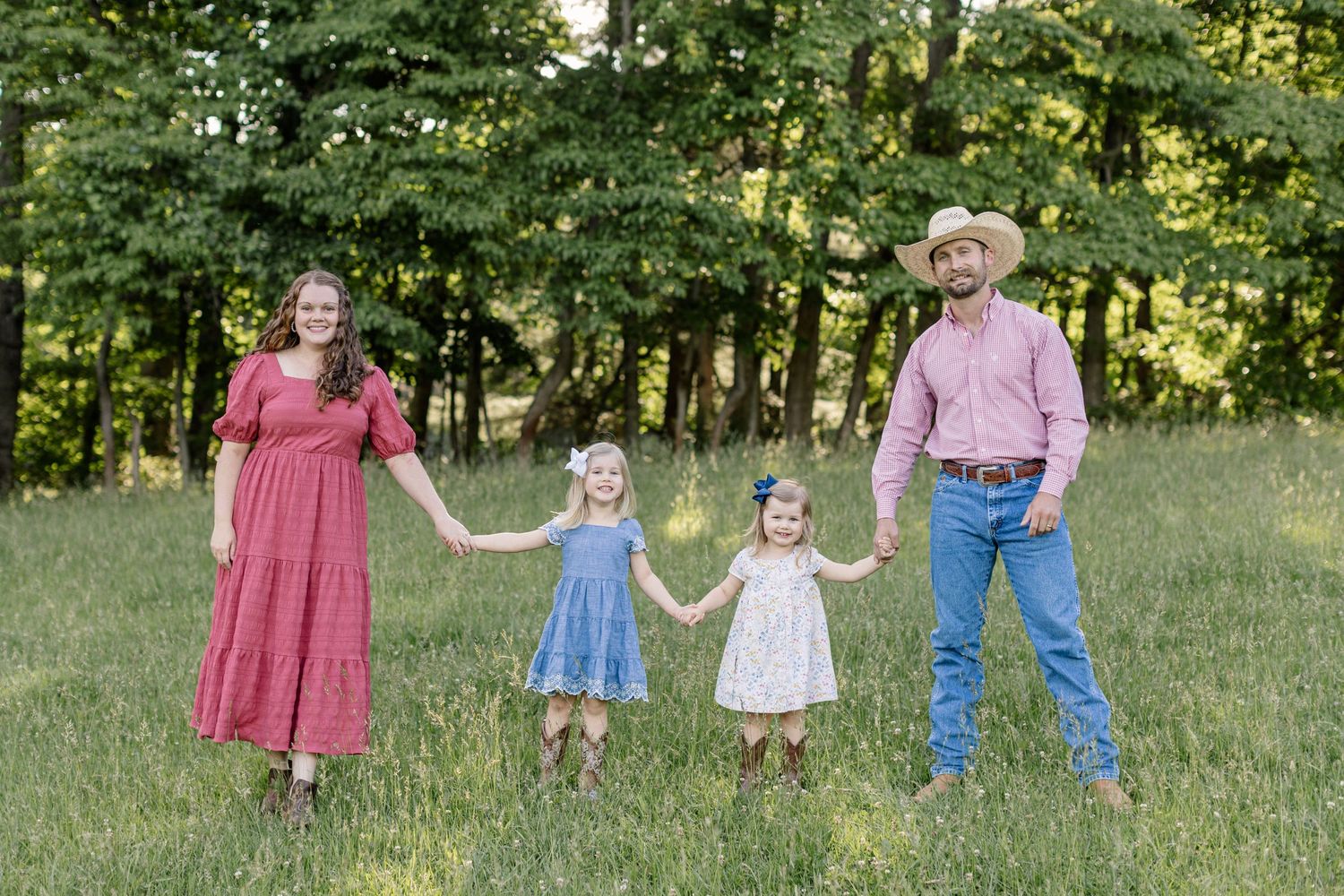 A family holds hands while standing together in a grassy field with trees in the background during golden hour.