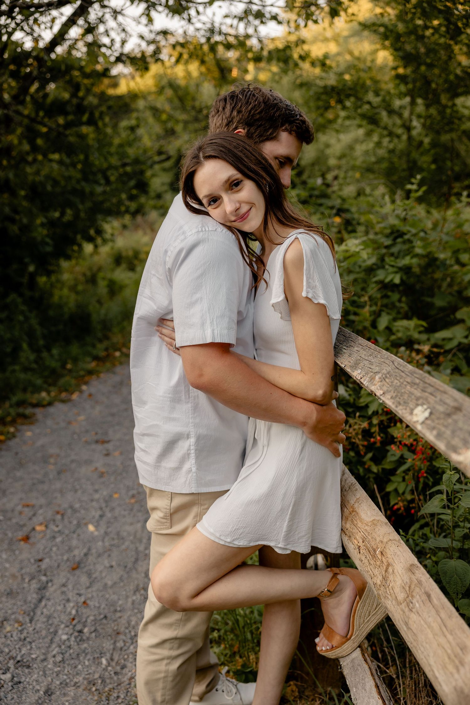 Couple leaning on wooden fence, woman resting head on fiancé’s shoulder, looking at camera – Pittsburgh engagement photo.