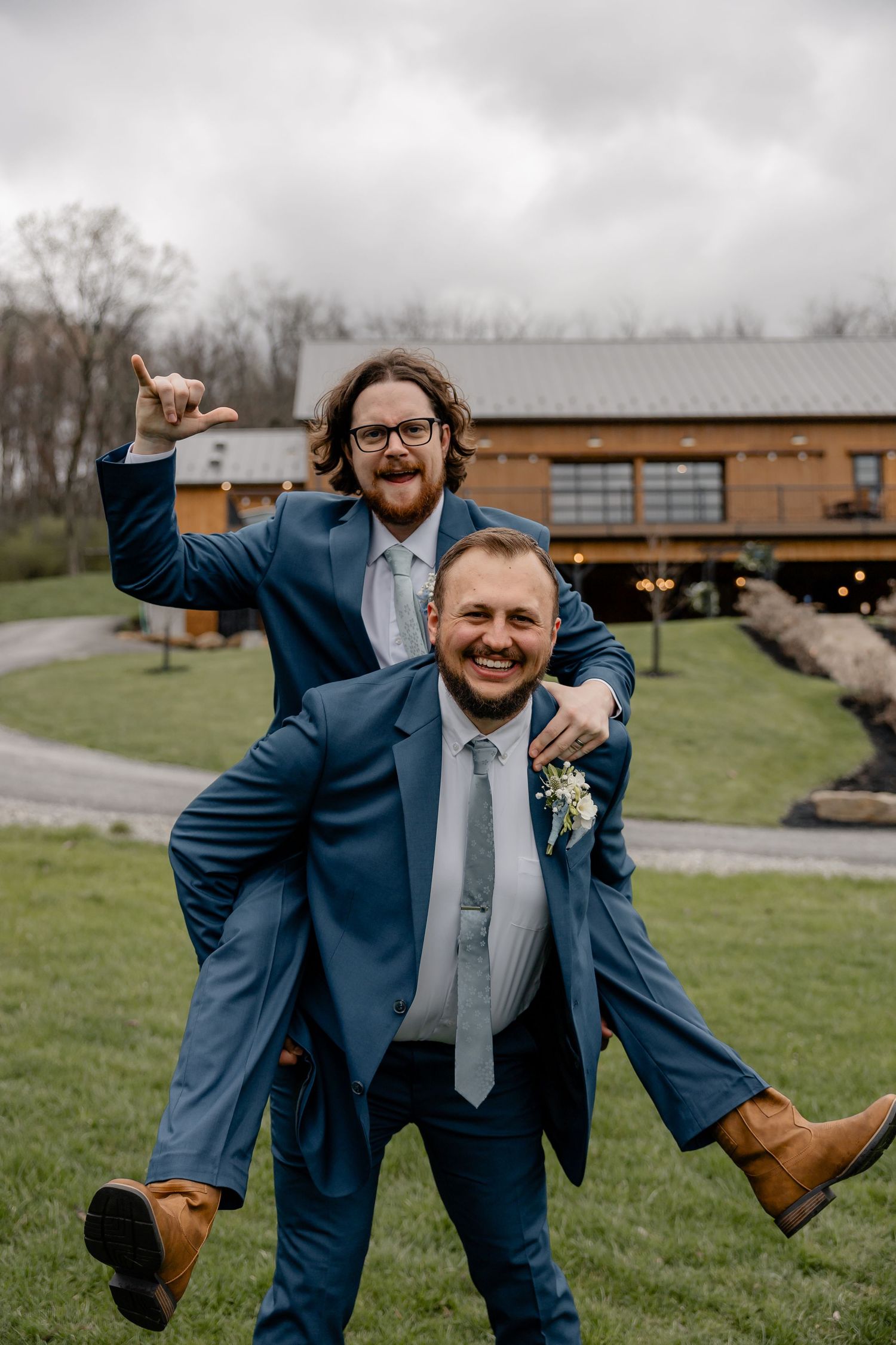 Best man playfully riding groom’s back, both smiling big with rustic lodge in background – fun Pittsburgh wedding moment.