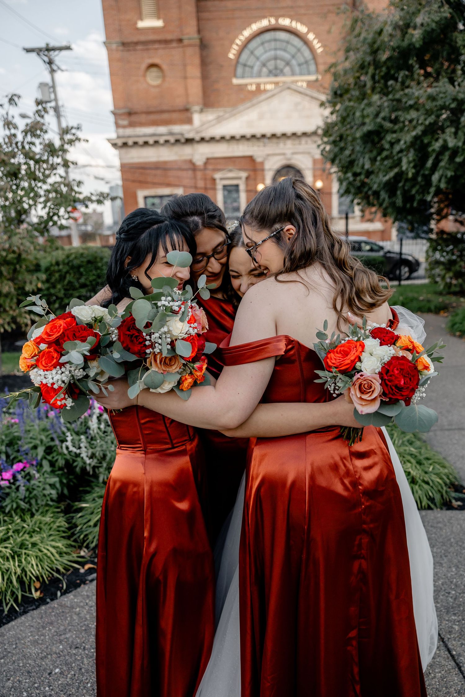 Bridesmaids in rust-colored dresses hugging with vibrant bouquets in front of Pittsburgh Grand Priory – natural light wedding photography by