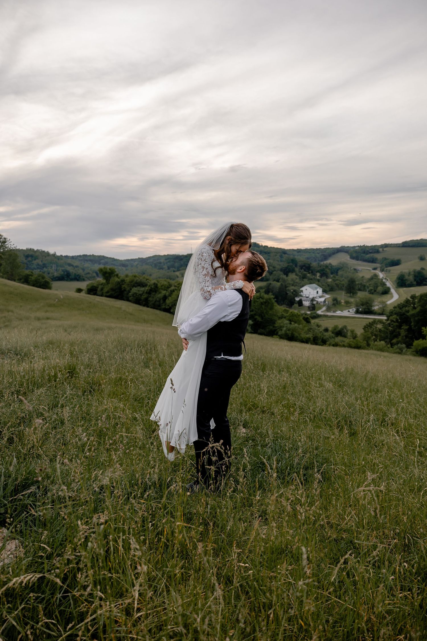 Groom holding bride in scenic sunset field, sharing kiss as her long wedding dress flows – Pittsburgh wedding photography.