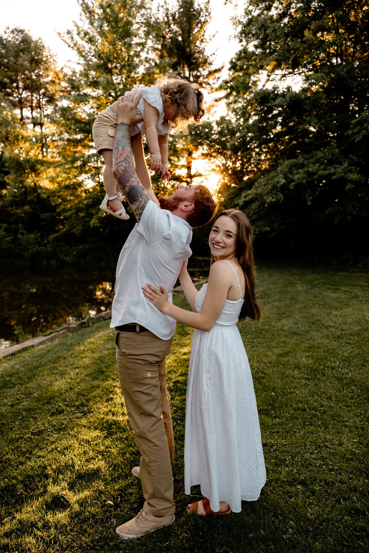 Man lifts toddler daughter in the air while woman holds him and laughs at camera – joyful family engagement photo at sunset in Pittsburgh.
