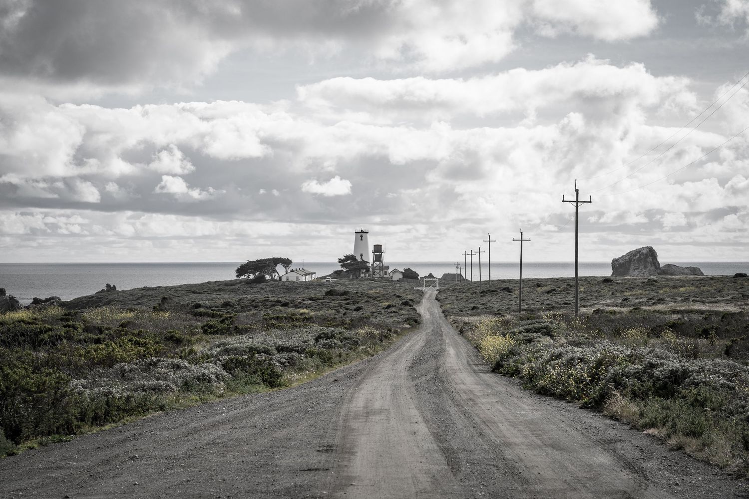 A black and white image of a lighthouse down a dirt road with fluffy clouds in Big Sur, CA.