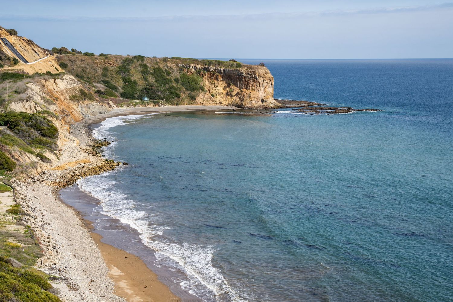 A cliff juts into the still turquoise waters of Palos Verdes. No one is on the small sandy beach.