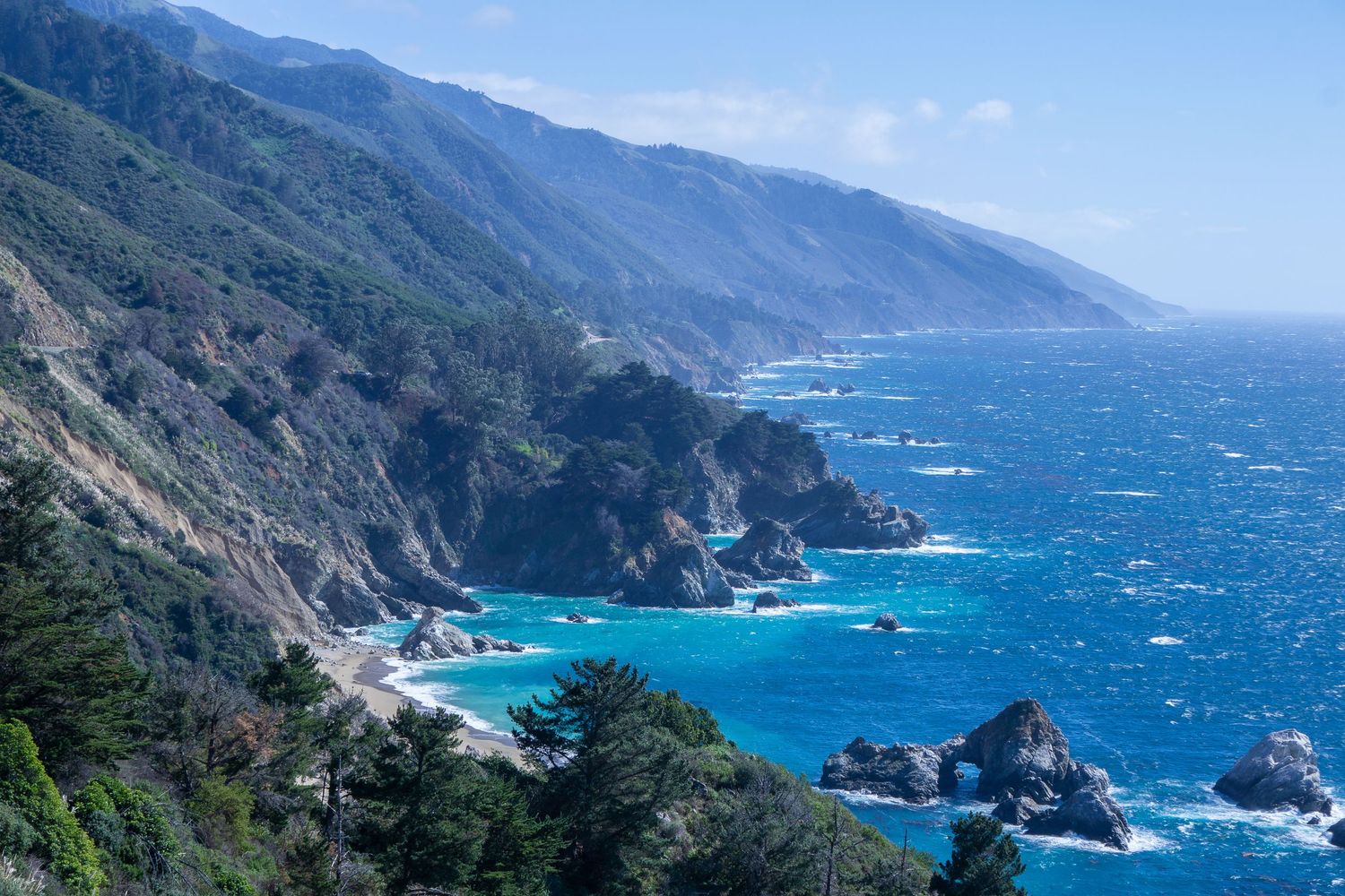 The cliffs of Big Sur sink into the Pacific Ocean on a windy, blue sky day.