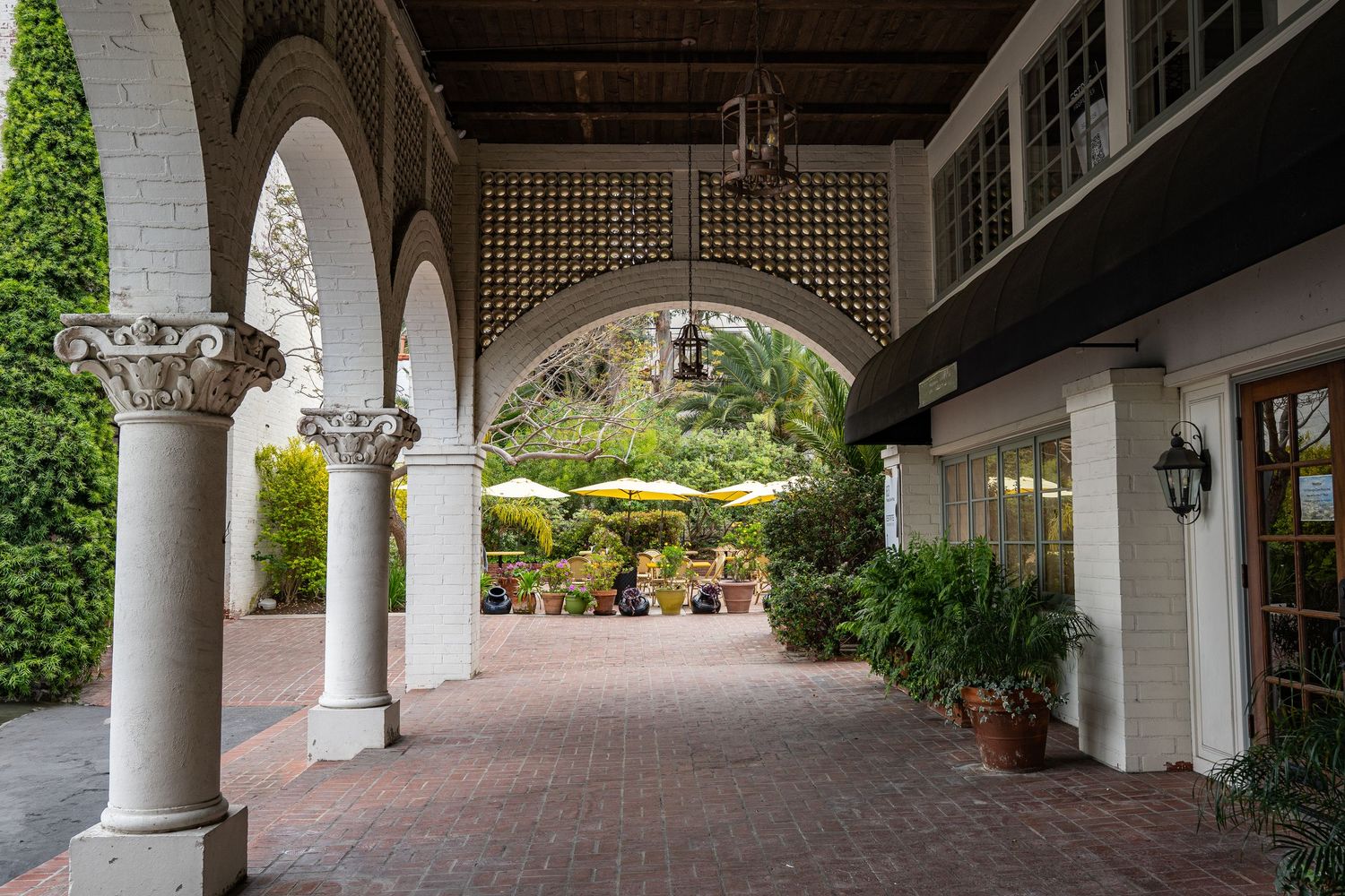 There are white archways along an old brick walkway. The ceiling is ornate and there is lots of greenery.