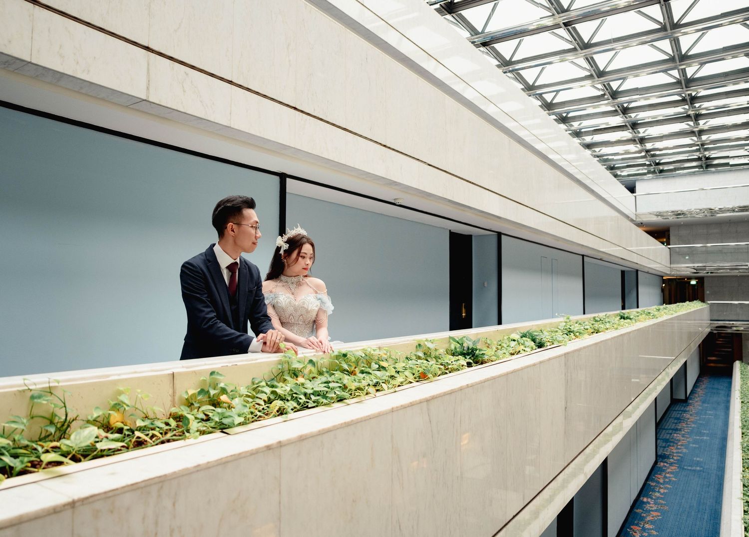 A couple in wedding attire stands together on a modern balcony with a built-in planter filled with green foliage.