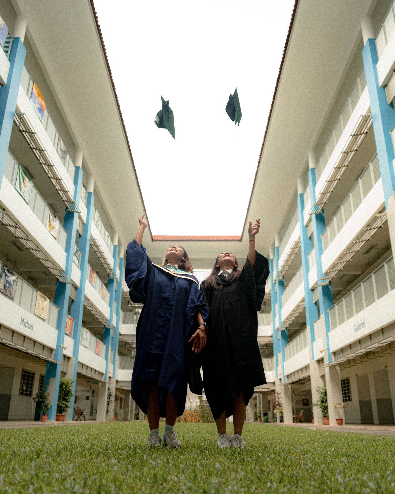 Two graduates in academic robes throw their caps in the air between school buildings.