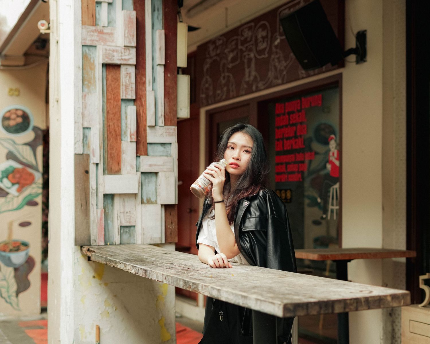 A person in a leather jacket leans on a cafe counter while drinking from a disposable cup with urban decor in the background.
