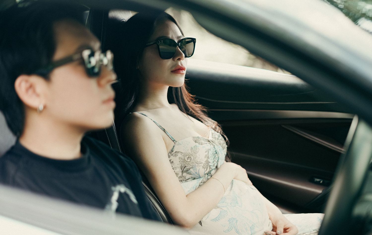Two passengers wearing sunglasses relax in a luxury white car, woman in floral dress seated in front.