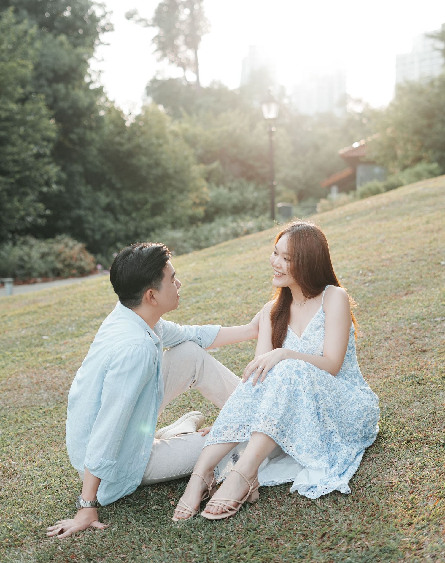 Couple sitting on green grass in a sunlit park, woman in floral dress and man in light blue shirt gazing at each other.