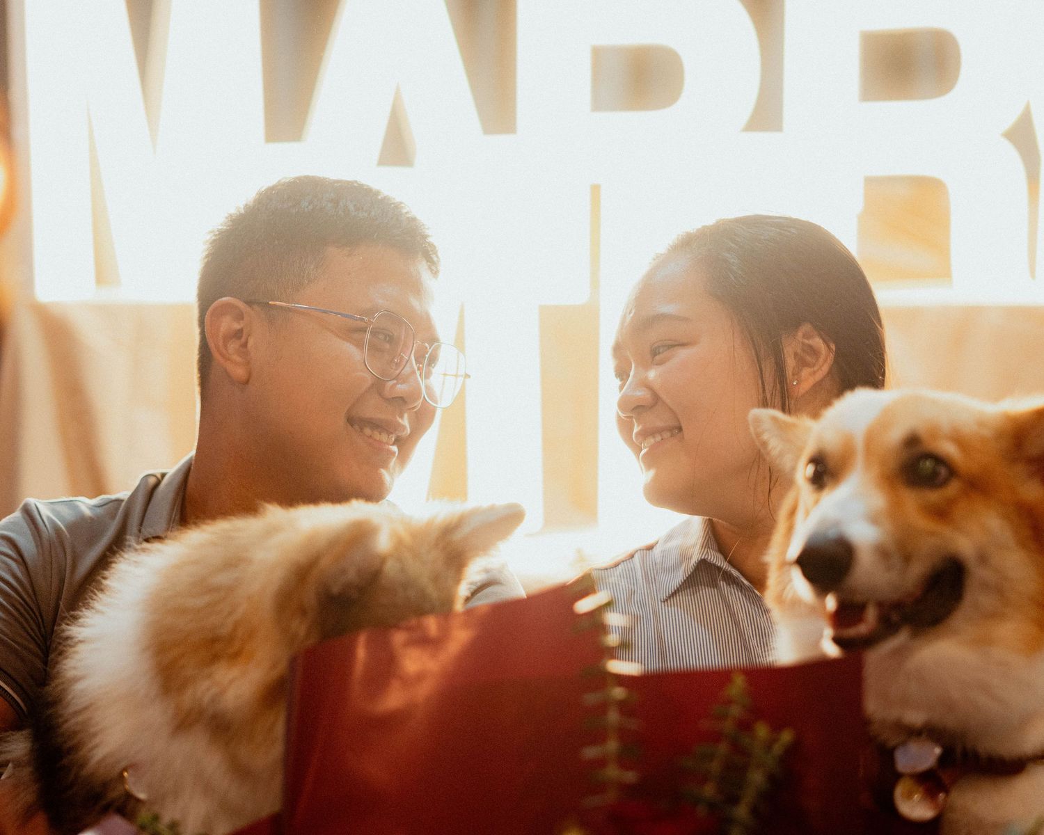 A couple sits on a couch with two adorable dogs while smiling at a bright MARR sign.