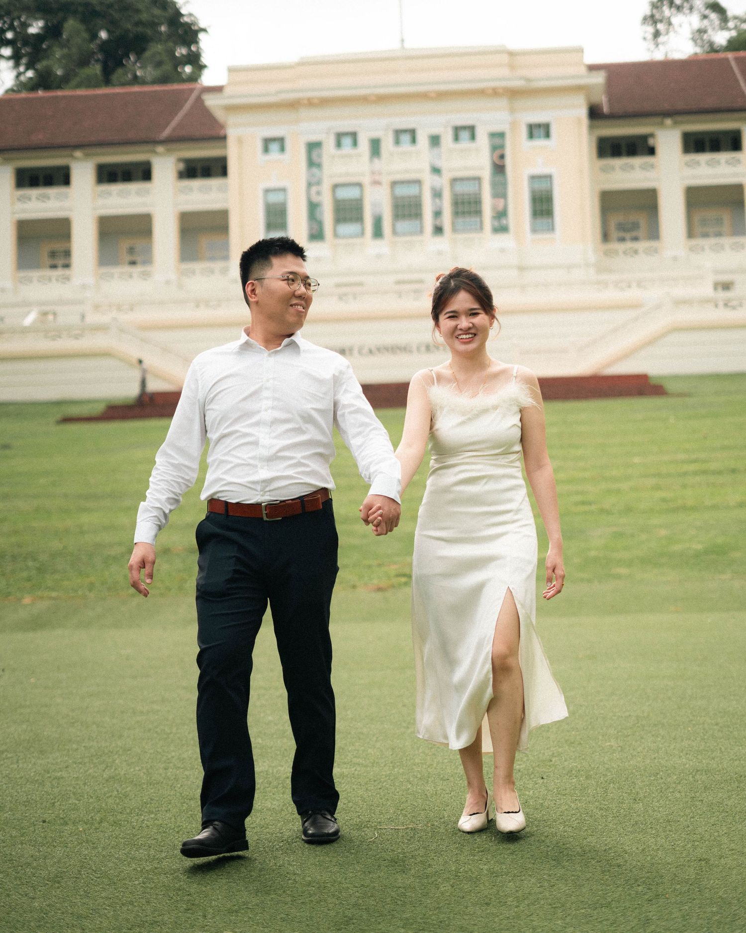 Couple strolling hand in hand on grass in front of a colonial-style building.