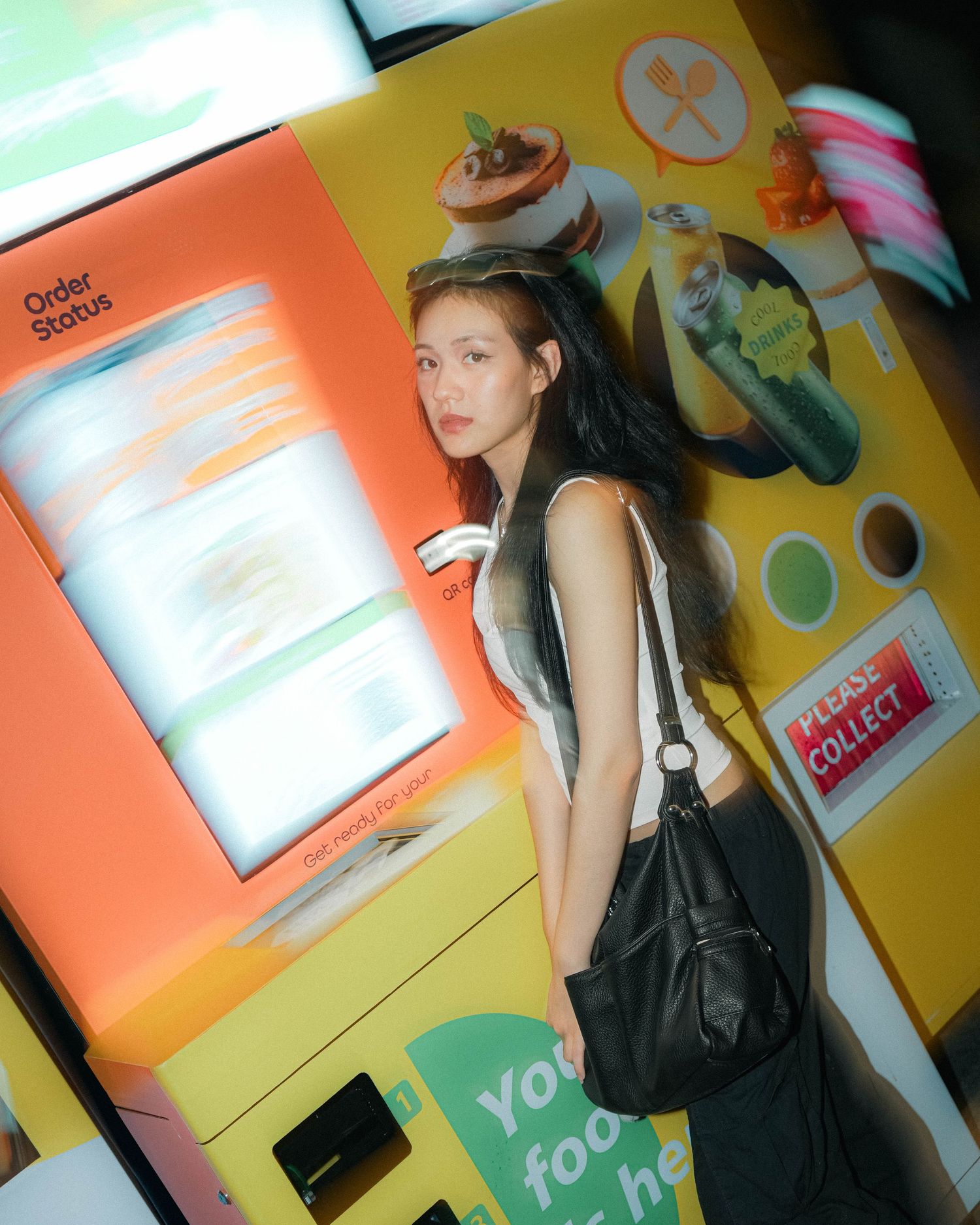 A person in a black outfit poses in front of a colorful vending machine wall with beverage advertisements.