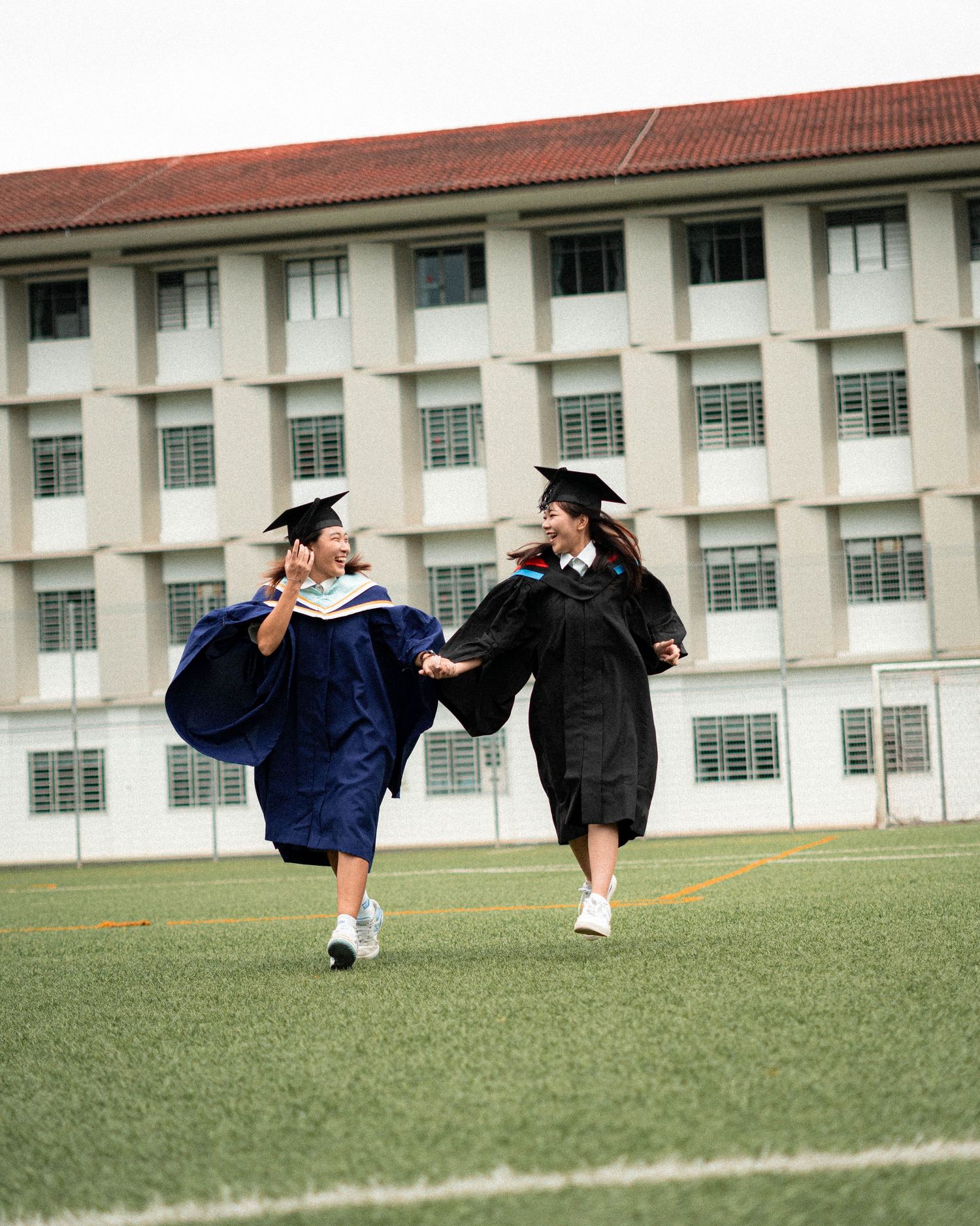 Two graduates in caps and gowns jump joyfully on a grass field in front of a university building.