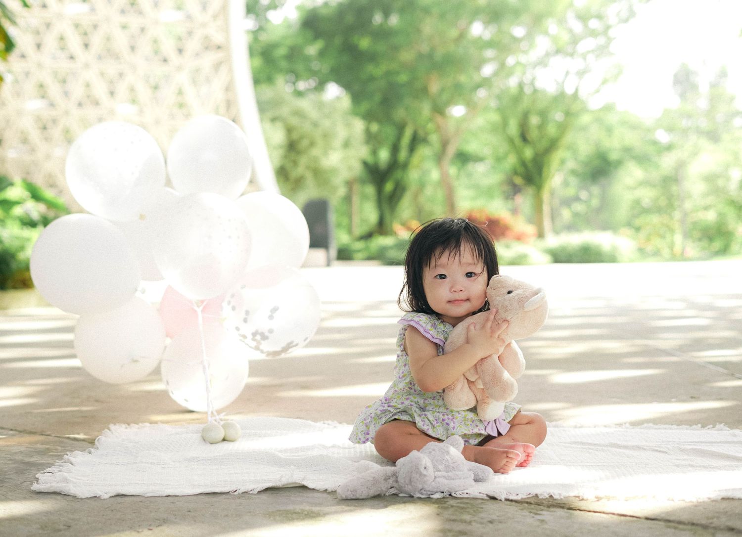 Small child sitting on white blanket with balloons in dreamy outdoor setting.