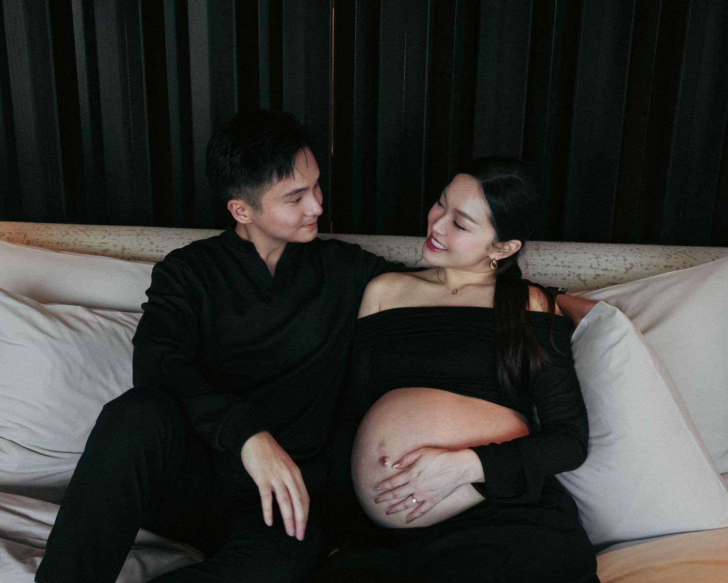 Couple in black attire sits together on white couch with pregnant woman cradling her baby bump.