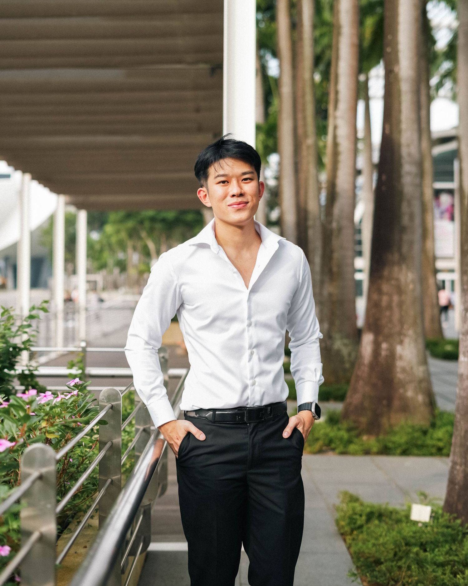Professional headshot with white dress shirt and black pants standing by palm trees and railing.