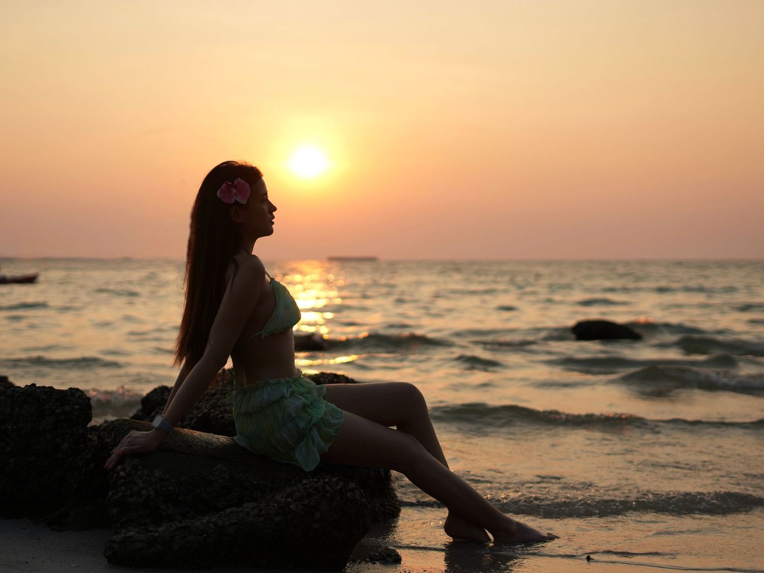 A silhouette sits on coastal rocks at sunset as golden light reflects off the ocean waves.