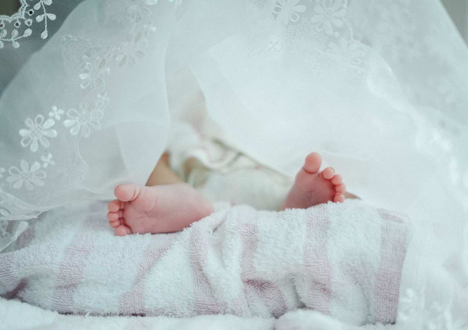 Tiny newborn baby feet resting on soft white lace and blanket in delicate lighting.