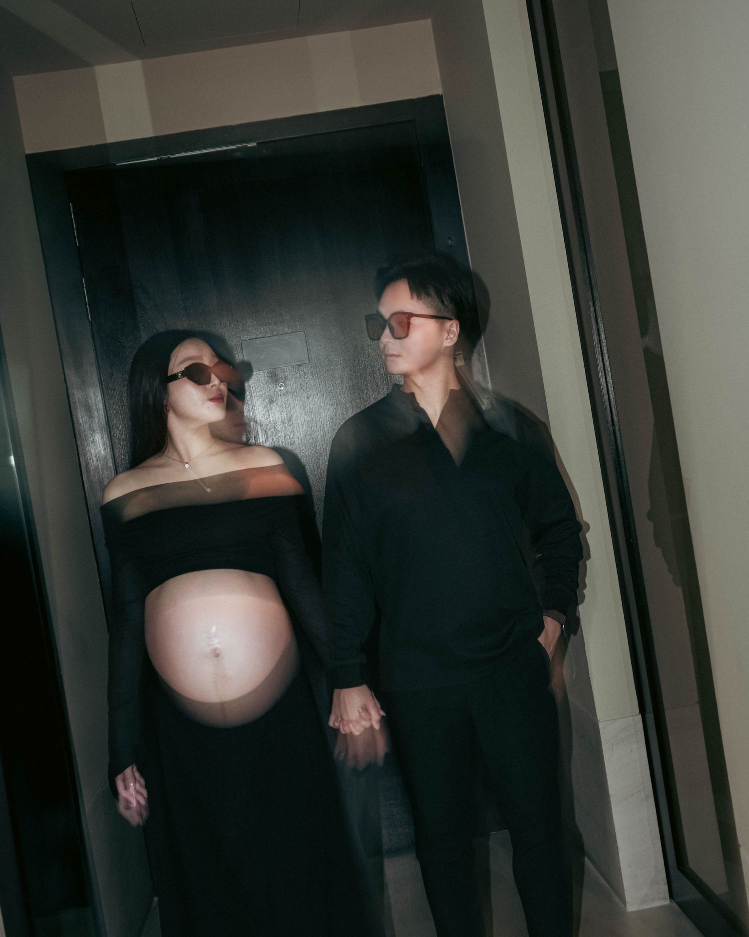 Artistic low light photo of a pregnant person in black attire with another person in dark clothing standing in a hallway.