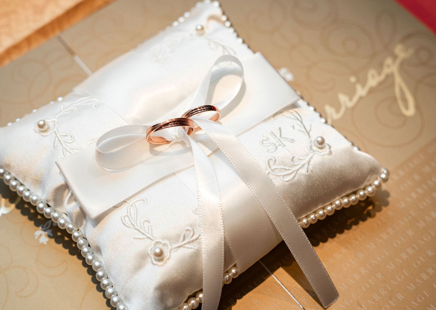 Two gold wedding rings resting on a white satin ring bearer pillow adorned with pearls and ribbon.