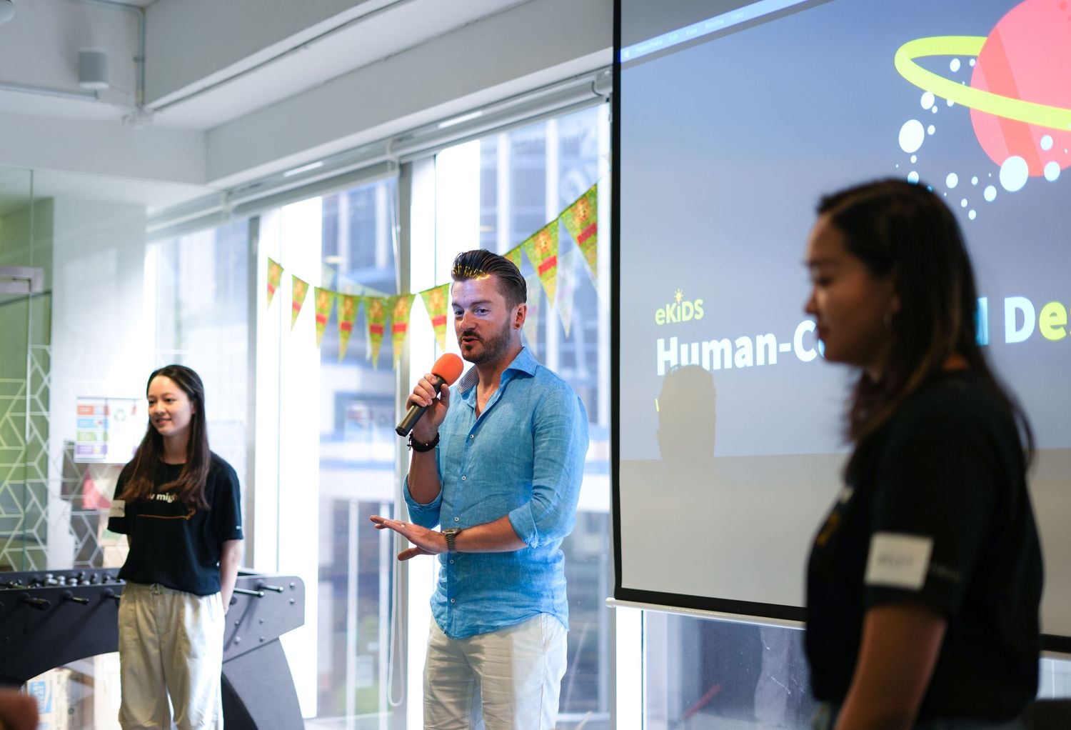 Presenters speaking at a business event with projection screen and colorful decorations in bright office space.