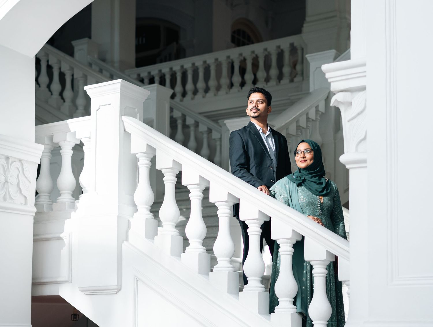 A couple in formal attire poses elegantly on a white ornate staircase in a grand building.