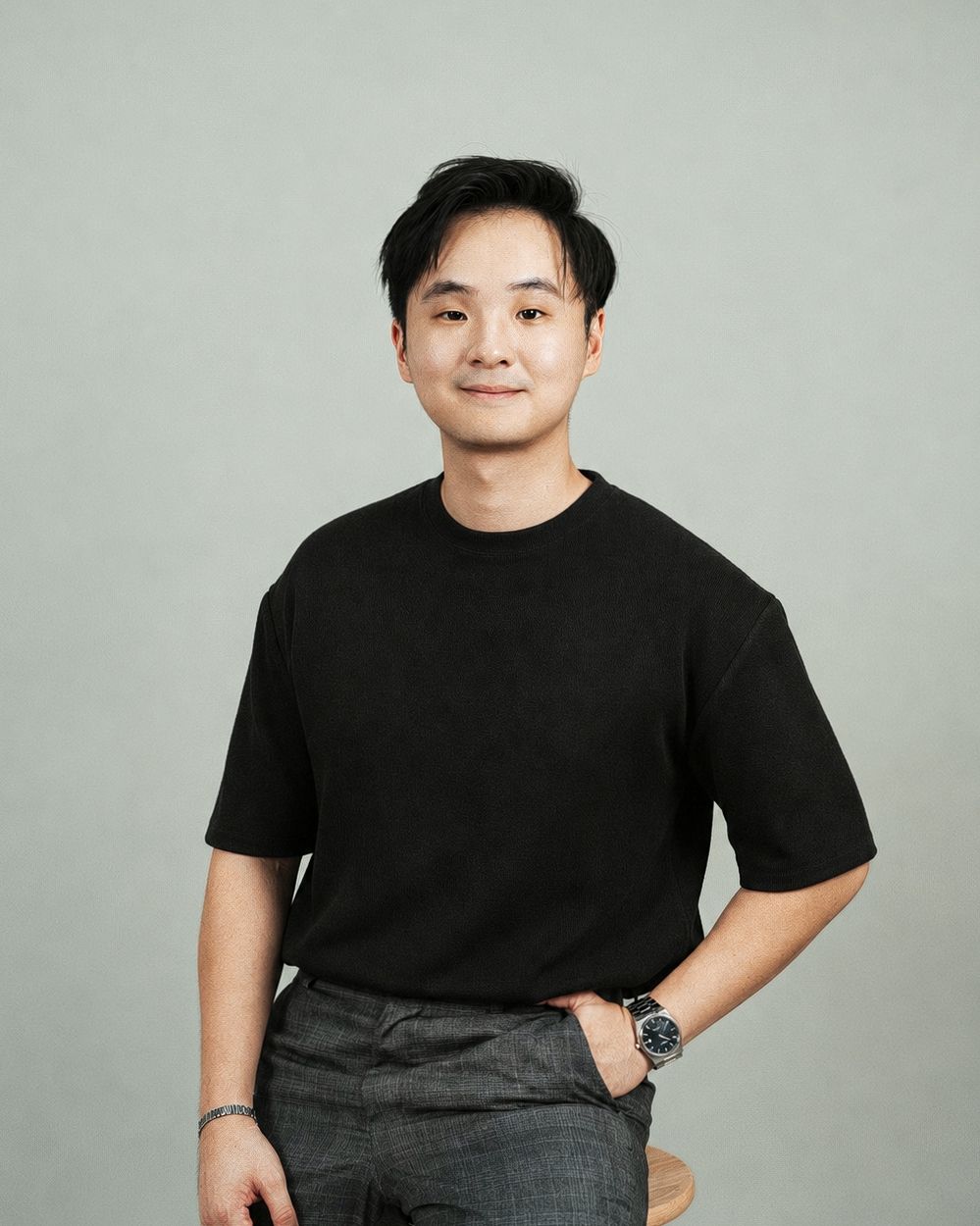 Young Asian man in black t-shirt and gray pants smiling confidently against a neutral gray background.