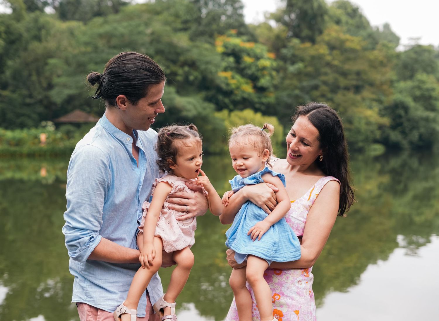 Two parents hold their young children near a scenic lake on a sunny day.