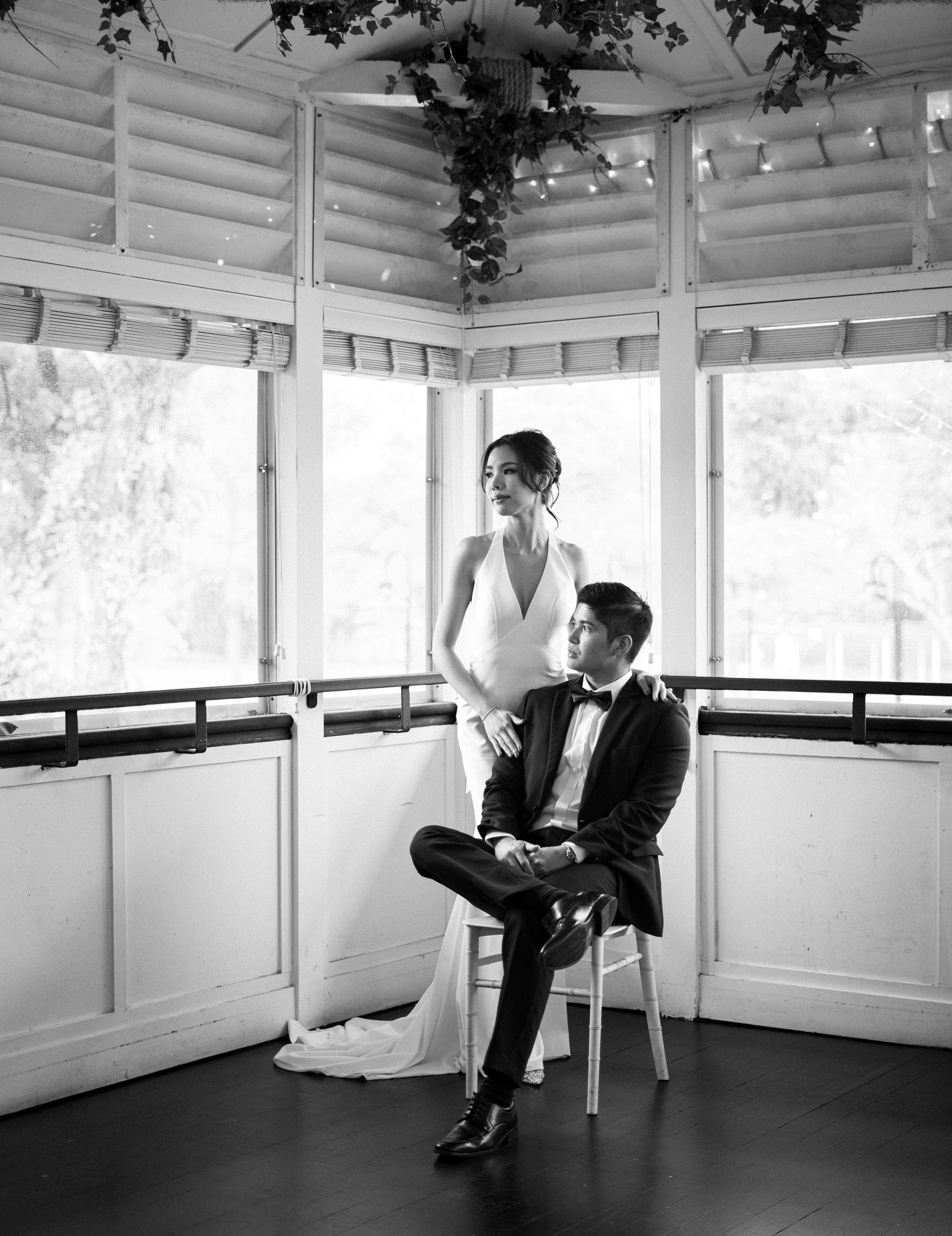 Black and white wedding photo of bride standing and groom seated in a sunlit pavilion with large windows.