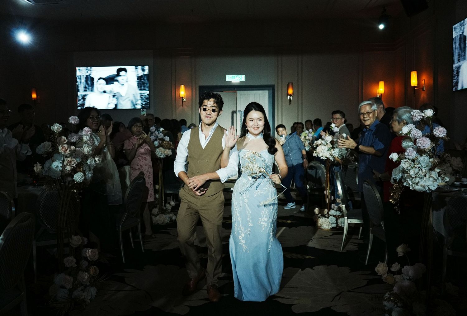 A couple walks down the aisle at their wedding reception, smiling joyfully, surrounded by guests and floral decorations.