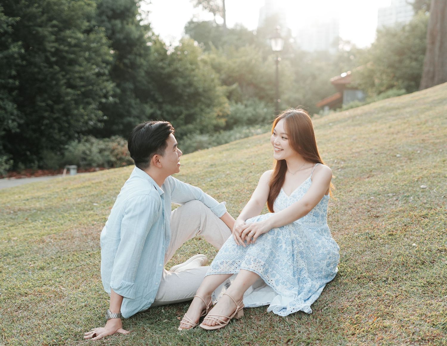 Couple sitting on green grass in a sunlit park, sharing a romantic moment in casual and floral outfits.