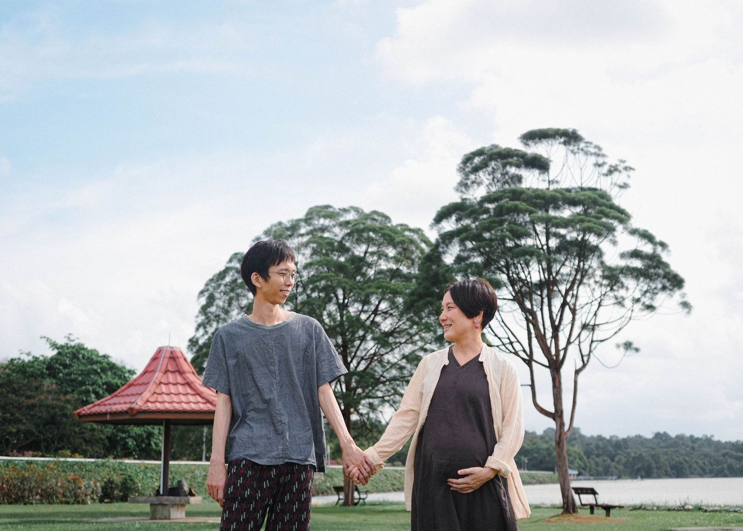 Two people holding hands while standing outdoors near a lake with tall trees and a gazebo in the background.