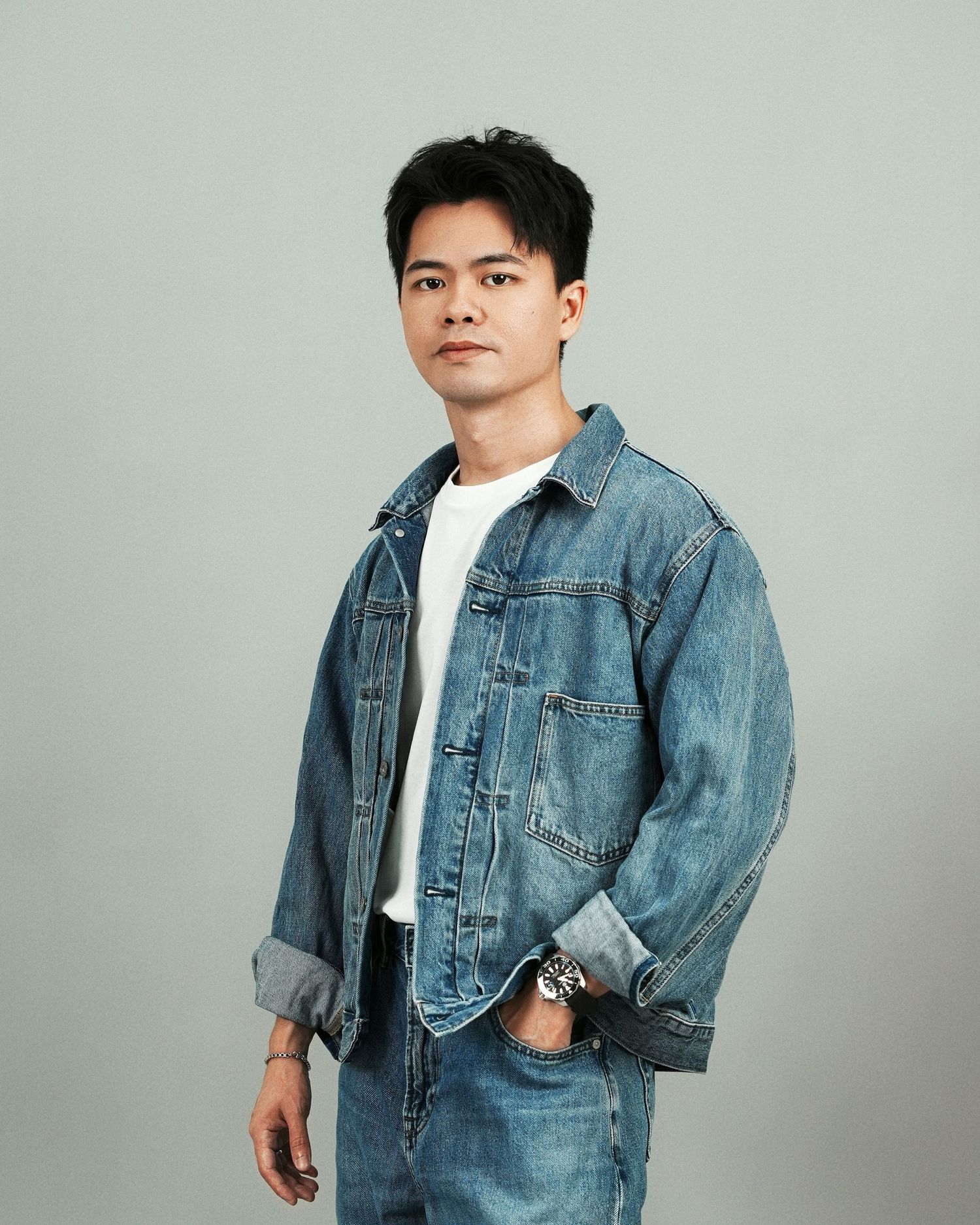 Young Asian man in denim jacket and jeans posing confidently against a light gray studio background.