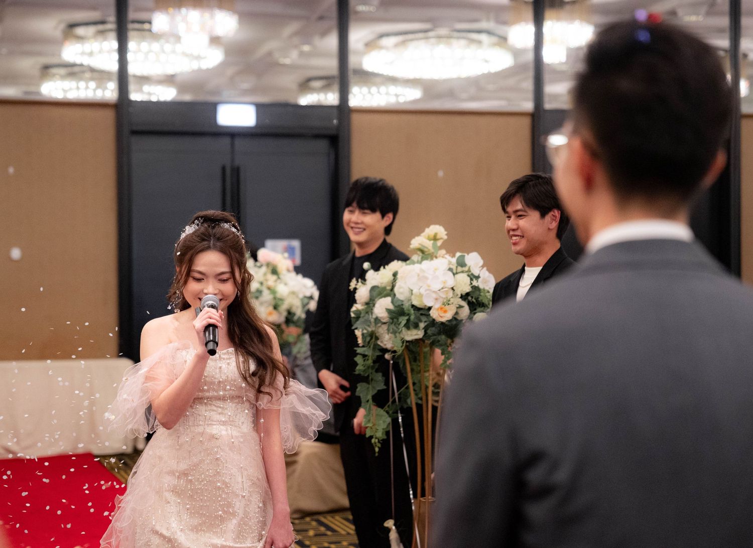 A person in a lace wedding dress speaks into a microphone while others stand nearby holding white floral bouquets.