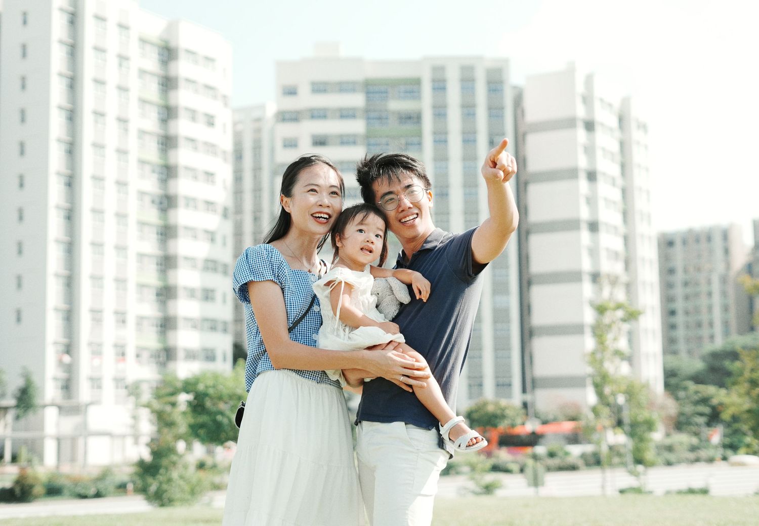 Happy Asian family with young child standing outdoors near modern apartment buildings, man pointing upward.