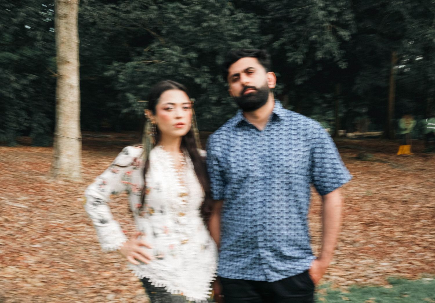 A couple stands together outdoors on a path covered with fallen leaves in a wooded area.