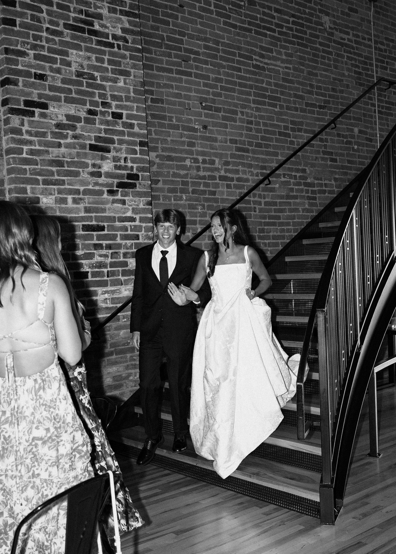 Black and white photo of a bride and groom descending a staircase at their wedding venue with brick walls.